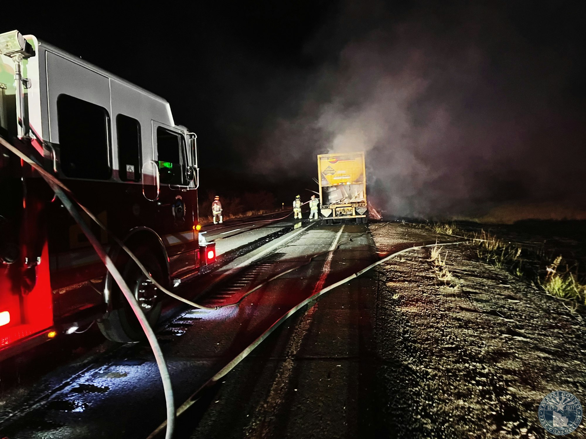 Firefighters next to a fire truck extinguishing a truck fire at night, with smoke visible.