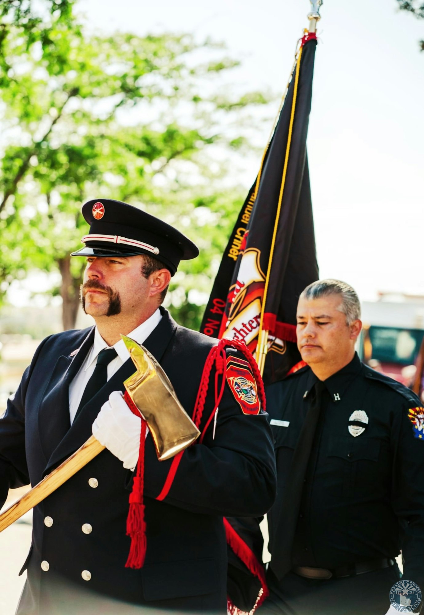 Two uniformed firefighters, one holding a ceremonial axe and flag, appear to be participating in an event or ceremony.