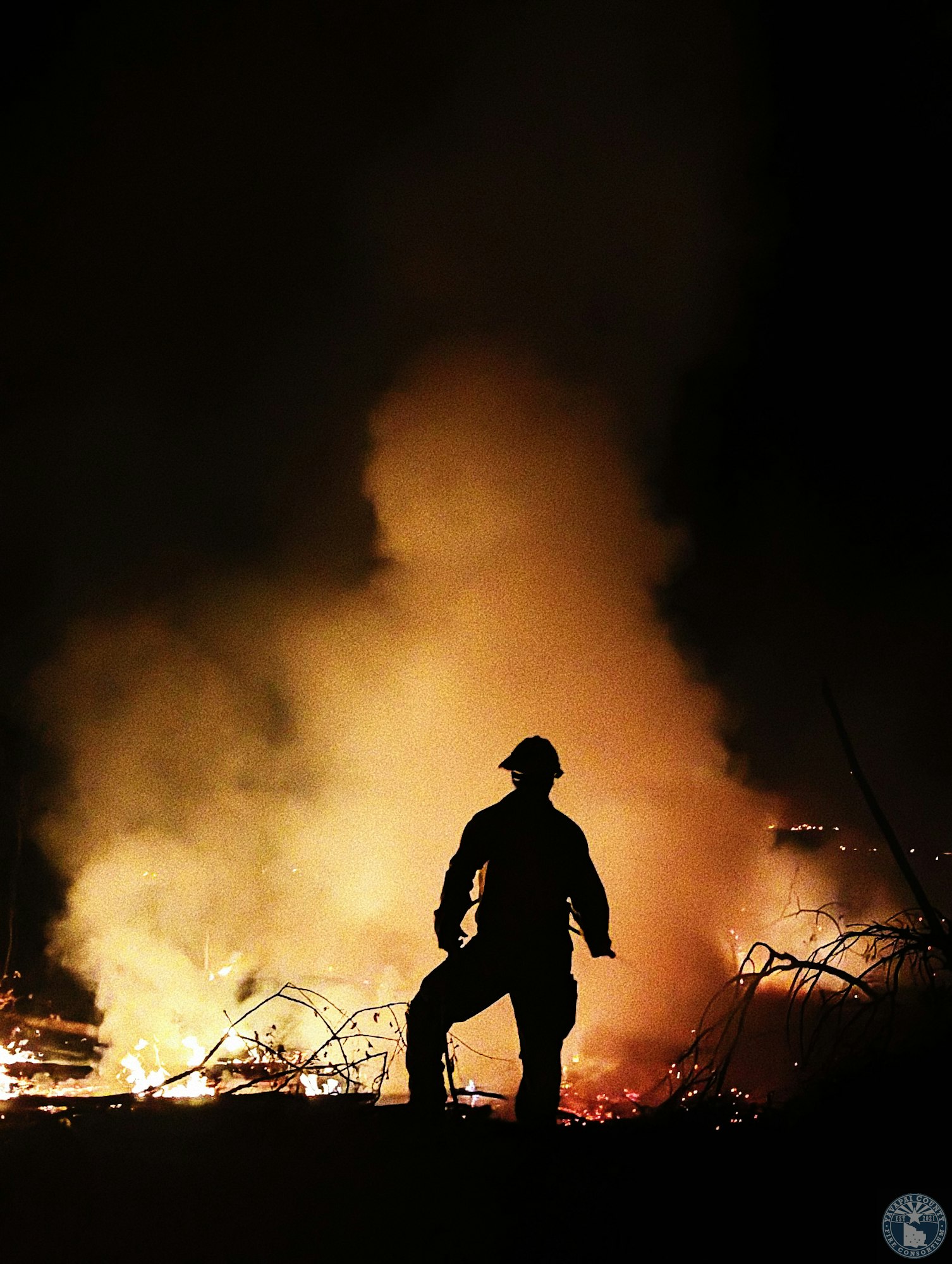 A silhouette of a person in protective gear, standing before a fire with smoke rising into the night sky.