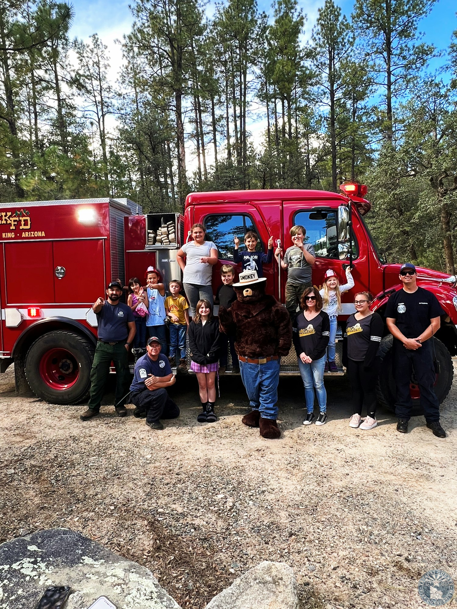 A group of people, including children and a bear mascot, pose in front of a red fire truck in a forest setting.