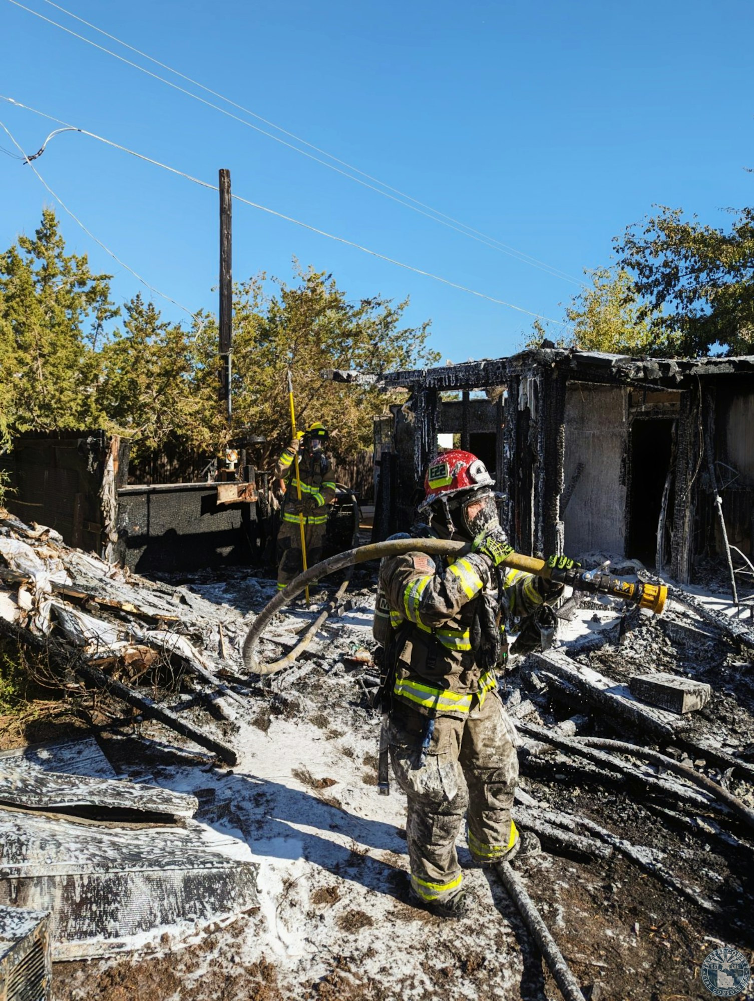 Firefighters in gear are extinguishing a burnt structure, surrounded by debris and foam, under a clear blue sky.