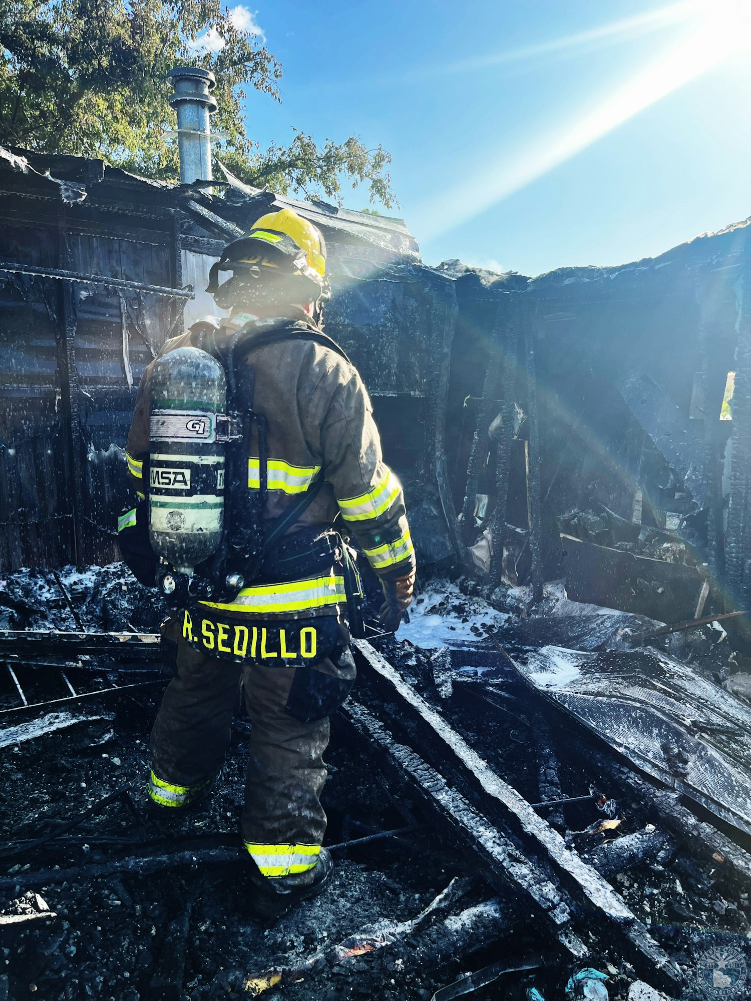 A firefighter in uniform stands amidst a charred, burned structure under sunlight.
