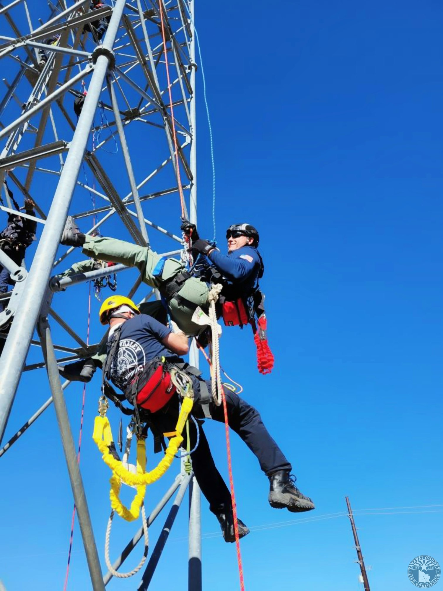 Two people in safety gear are climbing a metal tower, suspended by ropes against a clear blue sky.