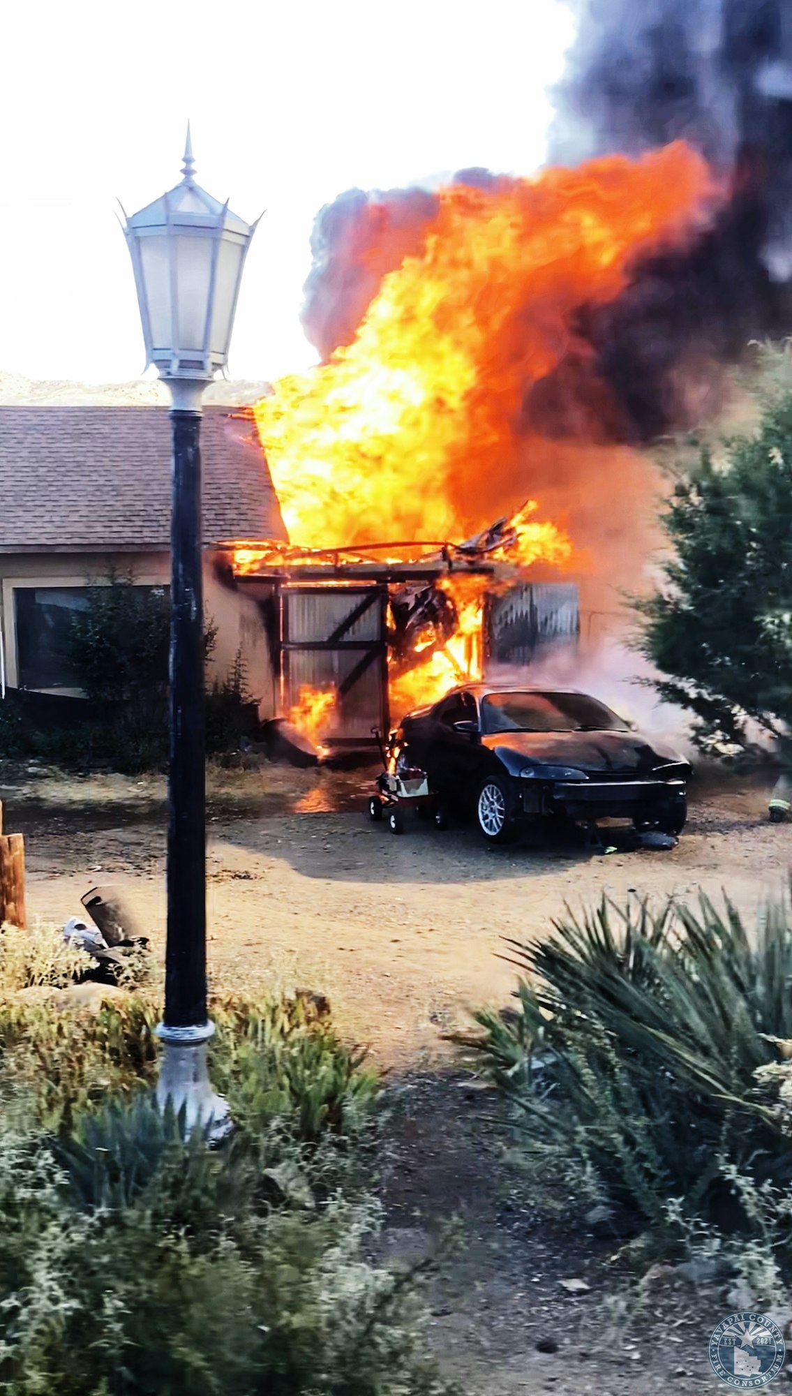 A house on fire with large flames and smoke, a car parked nearby, and a streetlamp in the foreground.