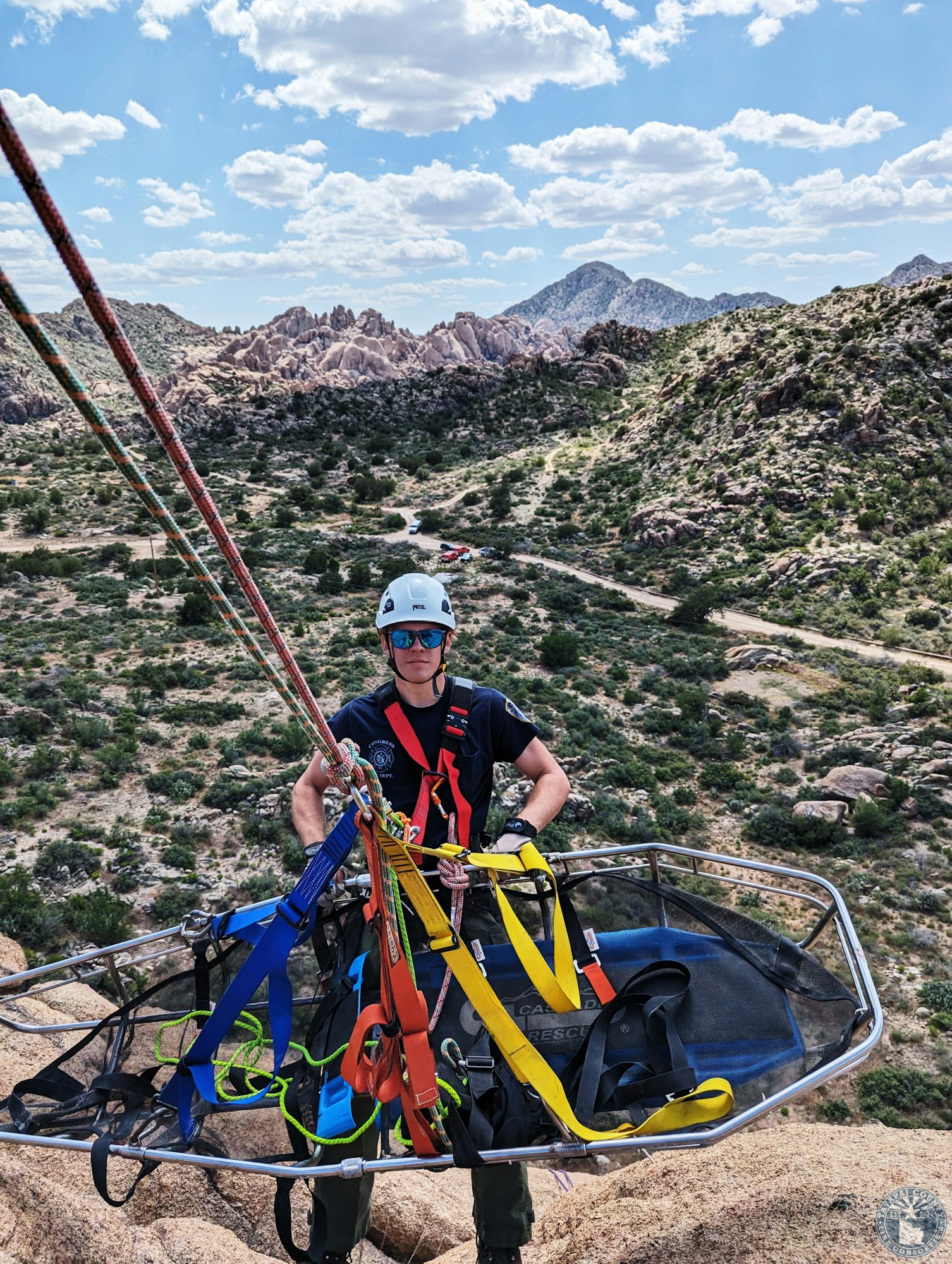 A person in a helmet and harness stands with rescue ropes and a stretcher atop rocky terrain under a blue sky.