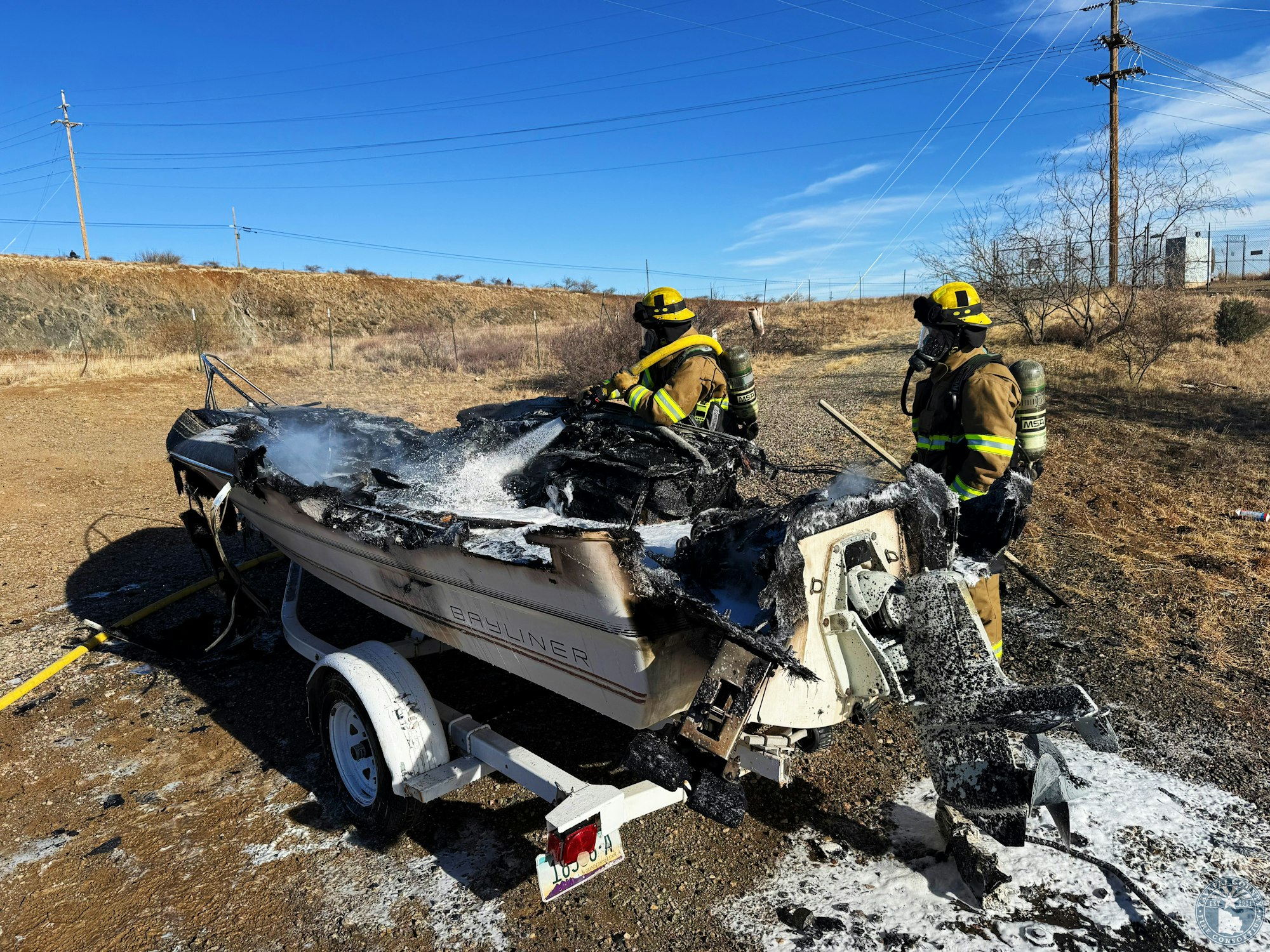 Firefighters extinguishing a charred boat on a trailer in a dry, outdoor area.
