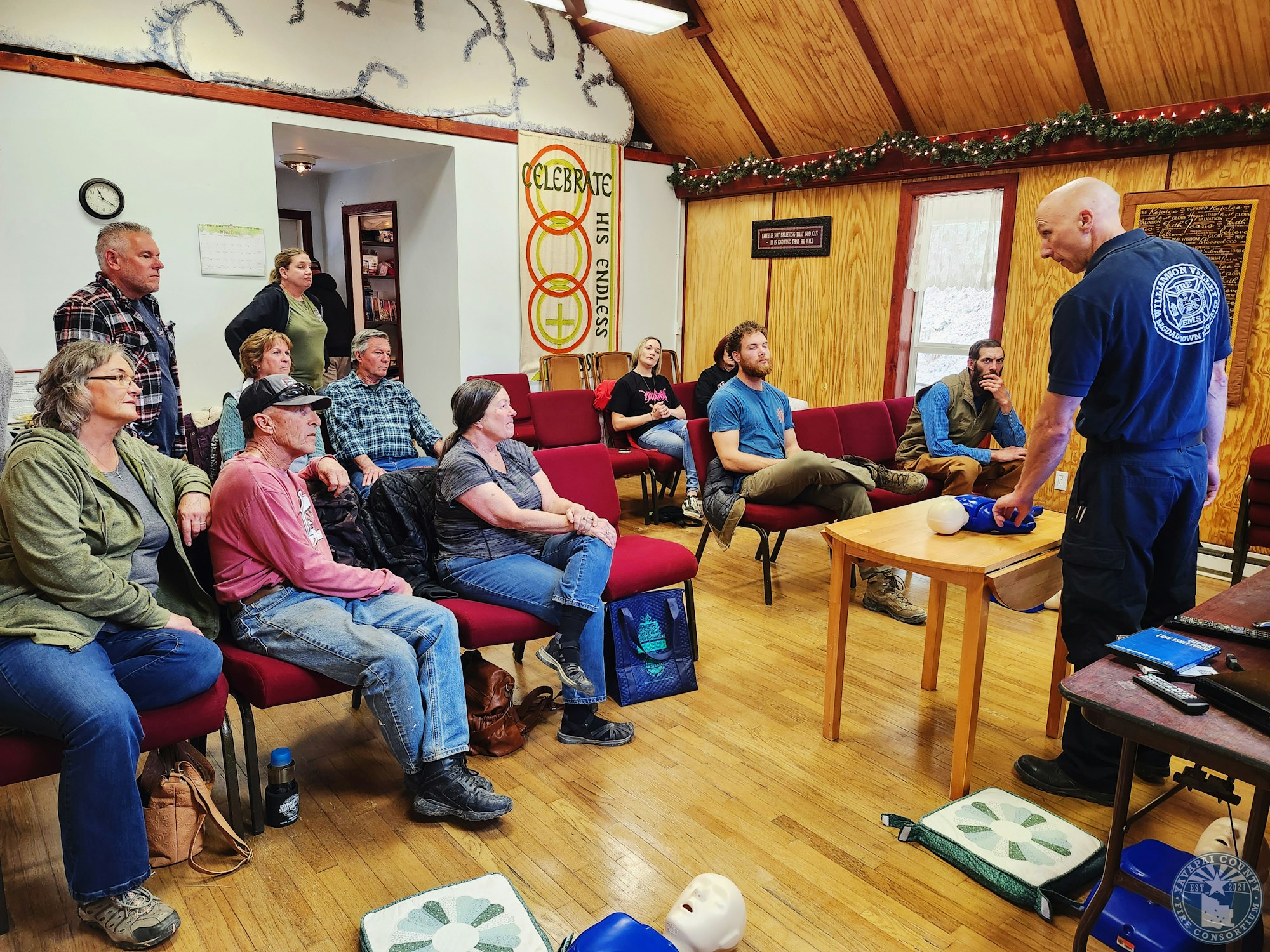 A group of people watch a first aid demonstration in a room with a CPR manikin on a table.