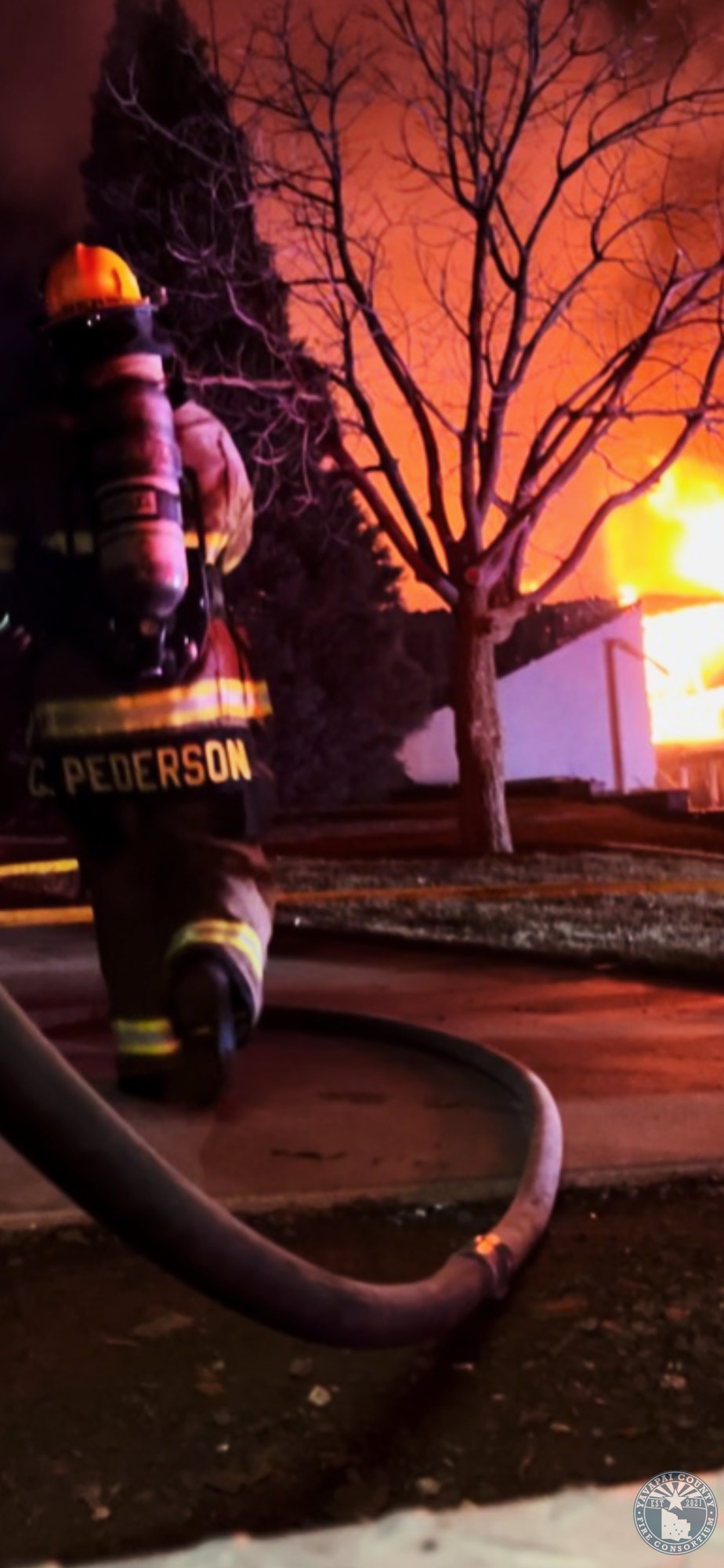Firefighter with hose approaches a large burning building, tree silhouettes against orange flames.