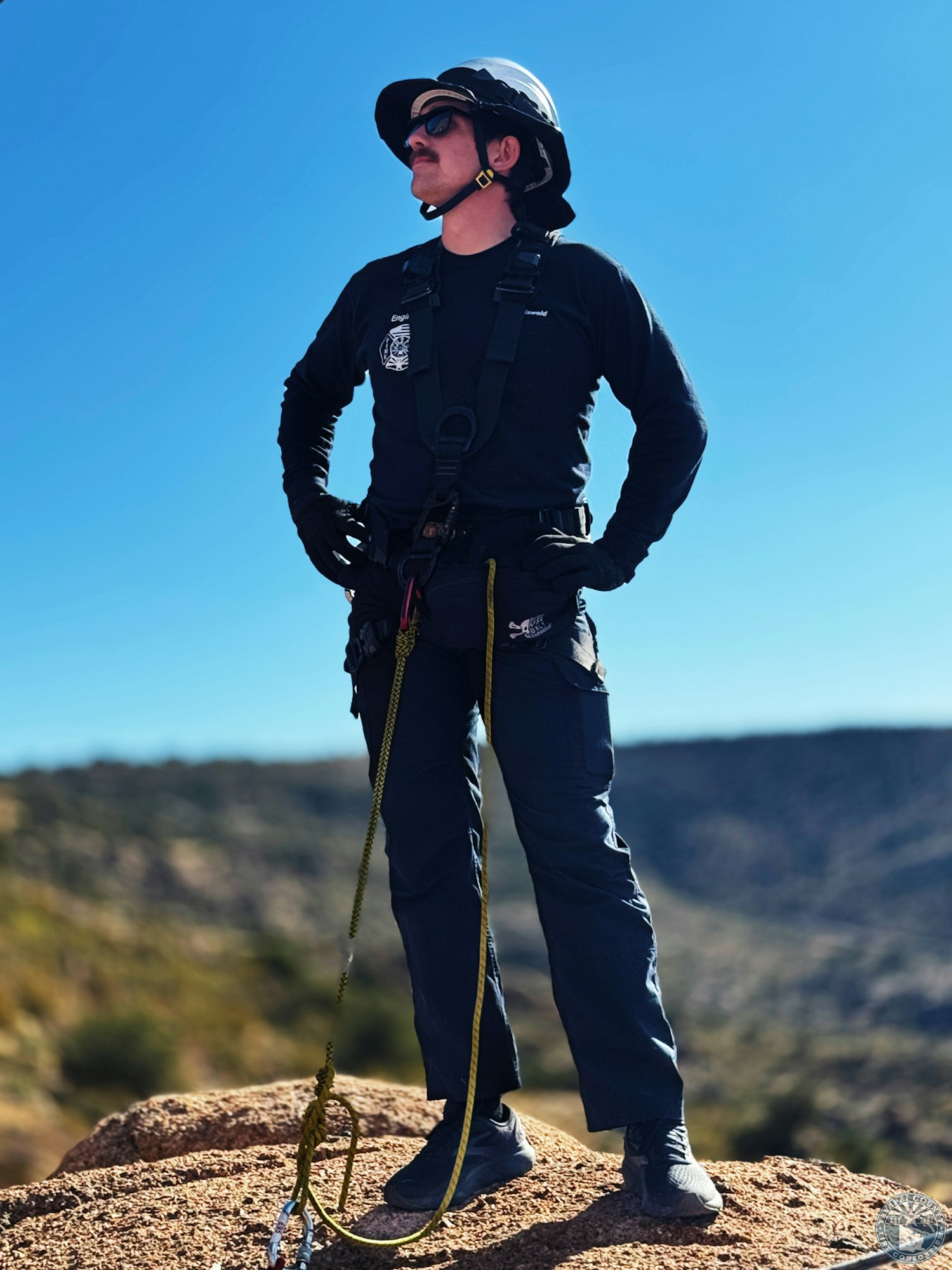 A person in outdoor gear and helmet, standing confidently on a rock with a rope, overlooking a mountainous landscape.