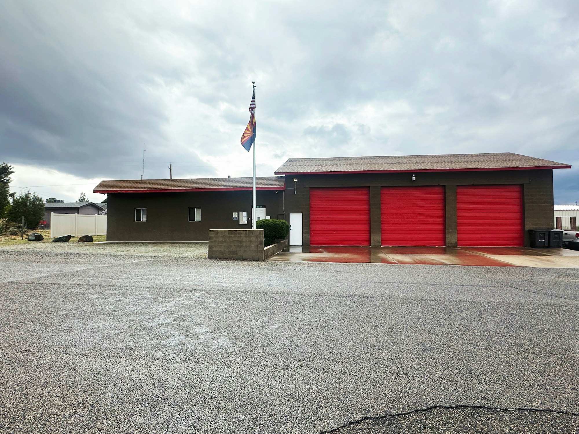 A building with three red garage doors, a flagpole with two flags, and an empty paved area in front under a cloudy sky.