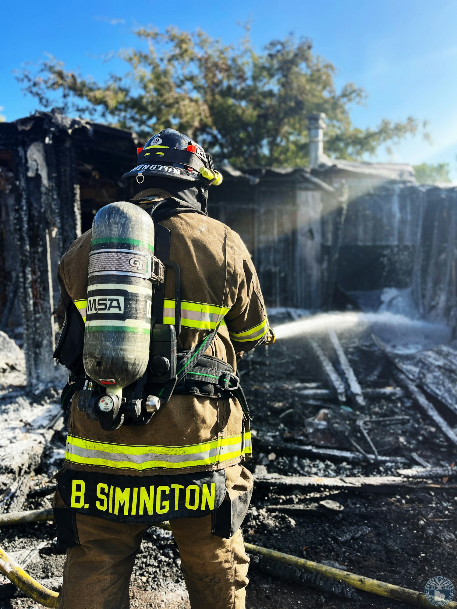 A firefighter in gear using a hose to extinguish a fire at a burnt structure, with trees in the background.