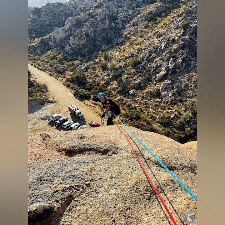 A person wearing a helmet is rock climbing with ropes, overlooking parked cars and rugged terrain below.