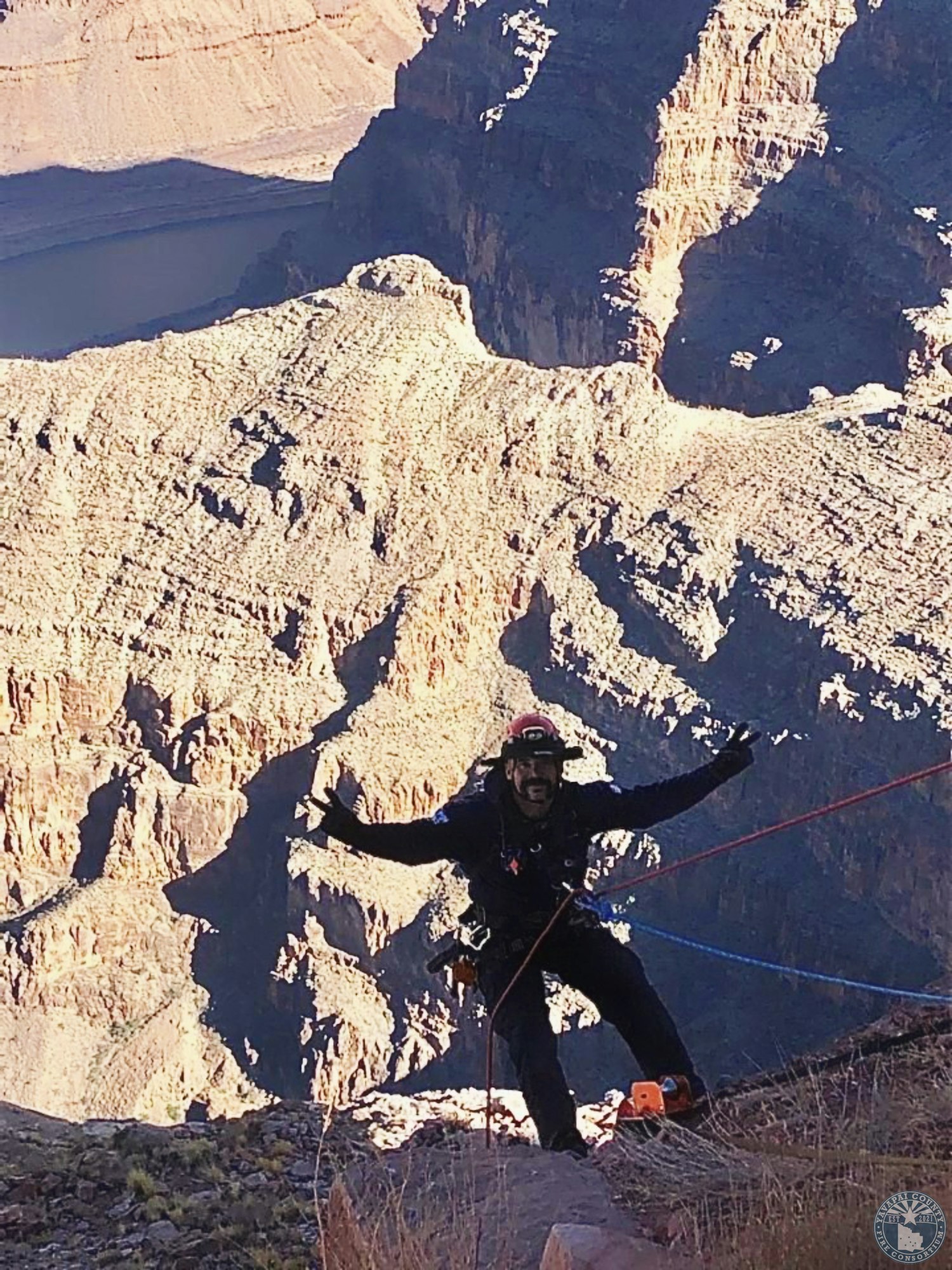 A climber with a harness smiles and gestures on a ledge with a canyon backdrop.