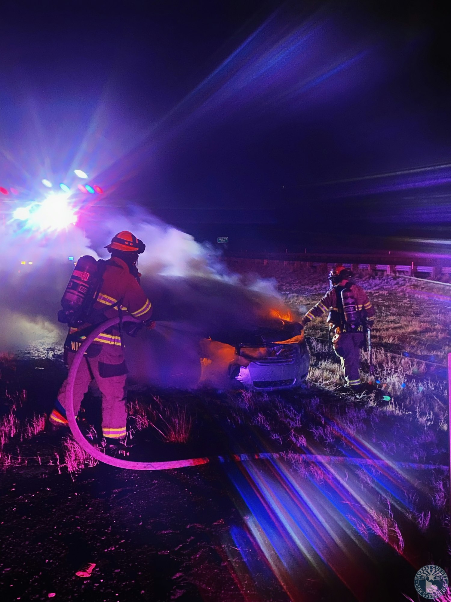 Firefighters extinguish a car fire at night, illuminated by emergency lights.