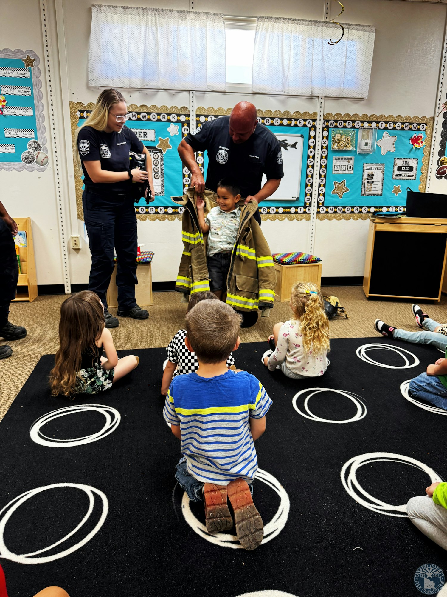 A young boy tries on a firefighter's coat with the help of two firefighters in a classroom, observed by sitting children.