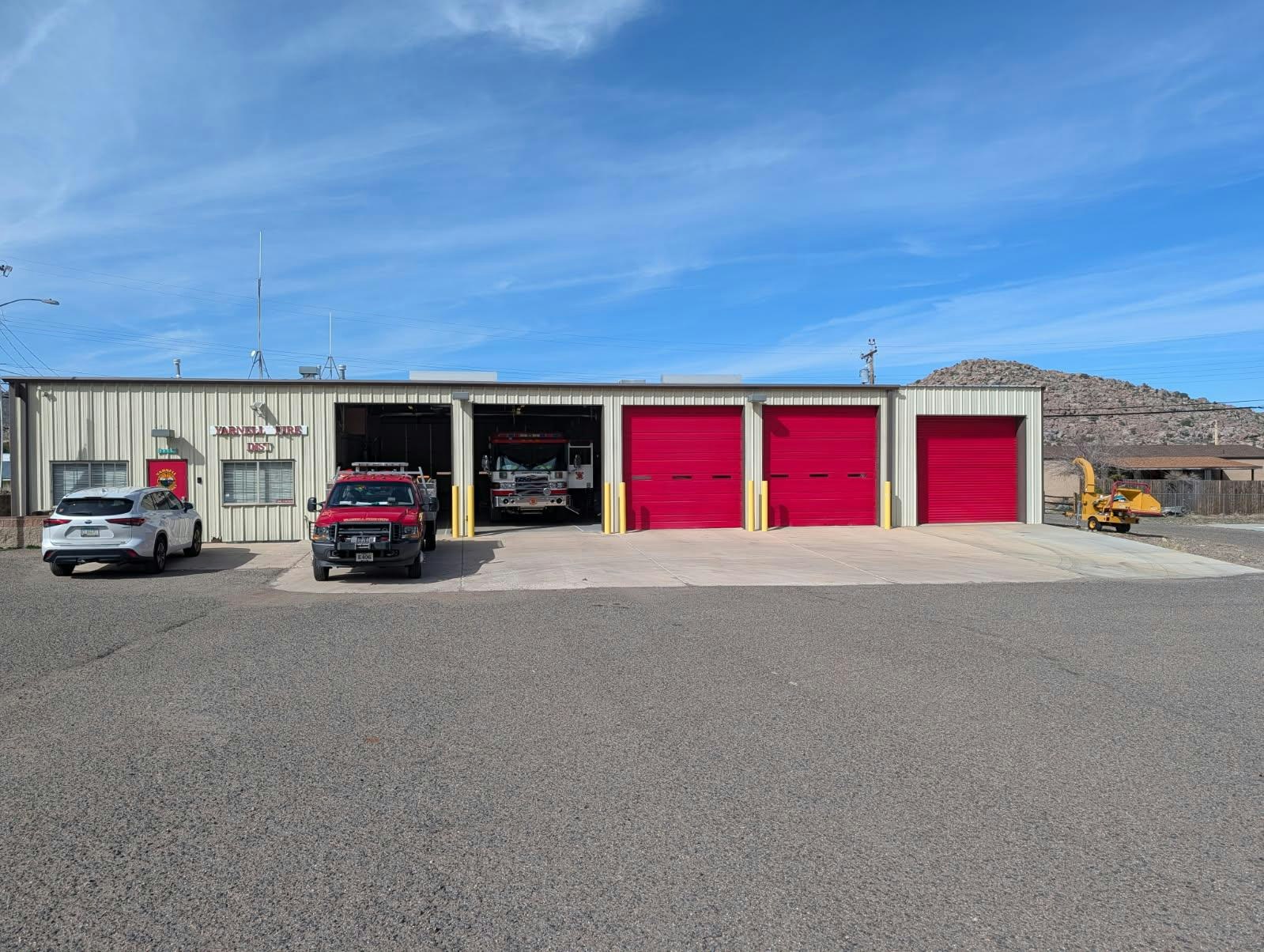 A fire station with red garage doors, a fire truck, two vehicles in the lot, and mountains in the background.