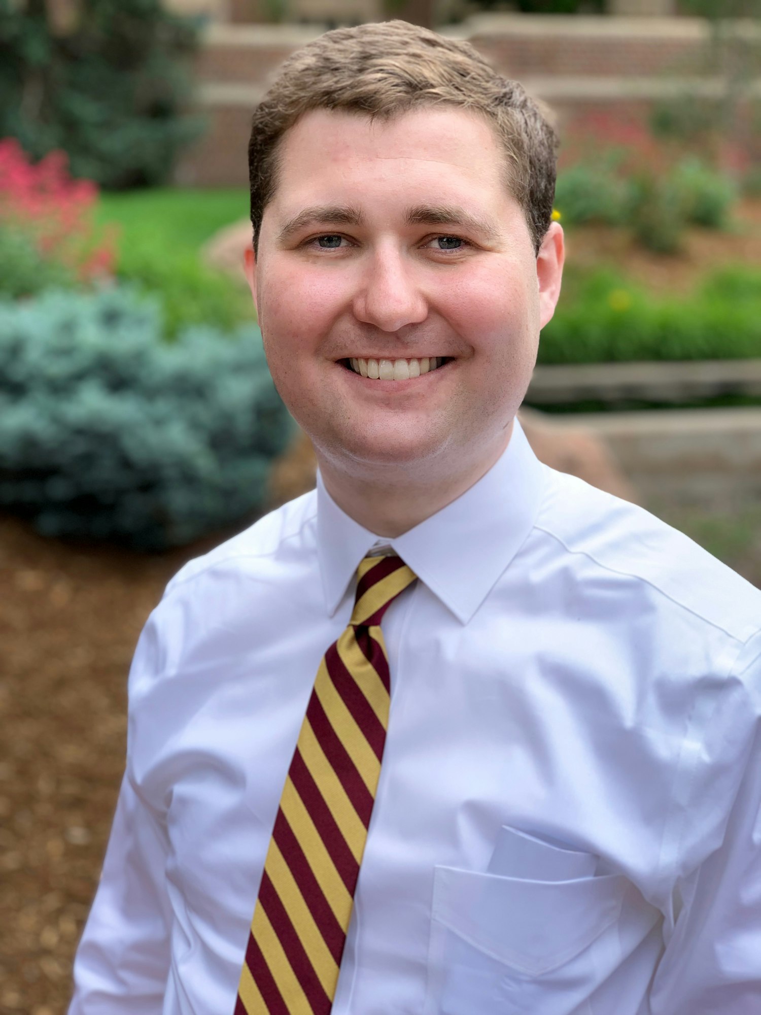 A smiling person wearing a white shirt and striped tie, outdoors with greenery in the background.