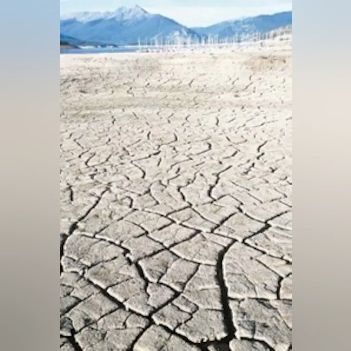 The image shows a dry, cracked landscape, suggesting drought conditions, with mountains and wind turbines in the background.