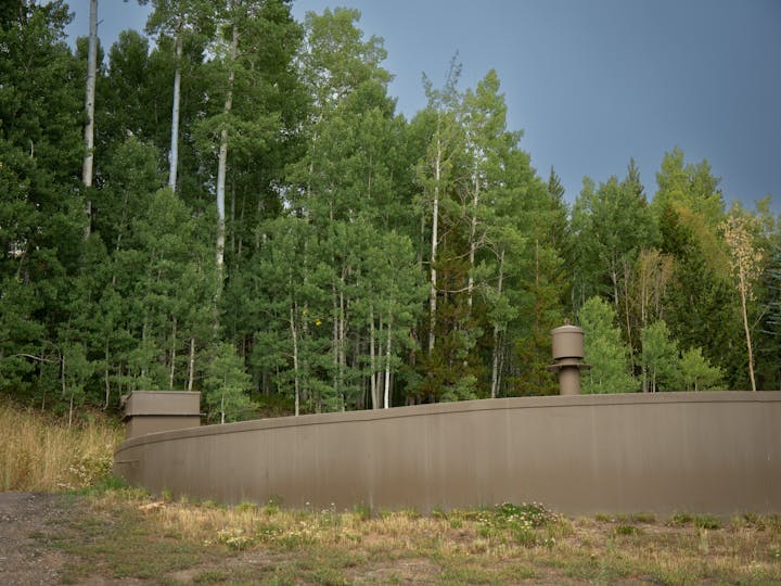 A curved, brown wall with two pipes in front of dense green trees and grass.