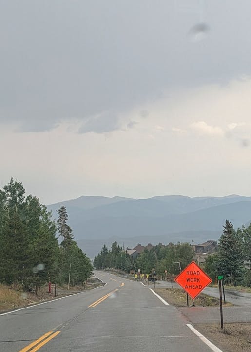 A winding road with "Road Work Ahead" sign, surrounded by trees and mountains under a cloudy sky.