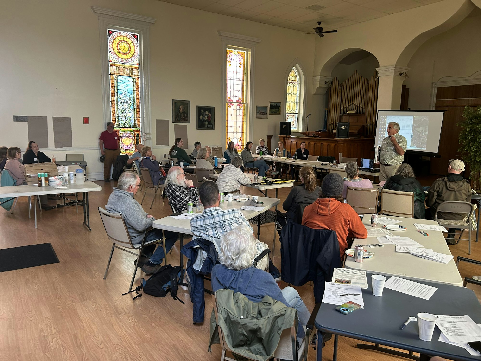 A group of people attends a meeting in a hall with stained glass windows, listening to a speaker presenting in front of a screen.