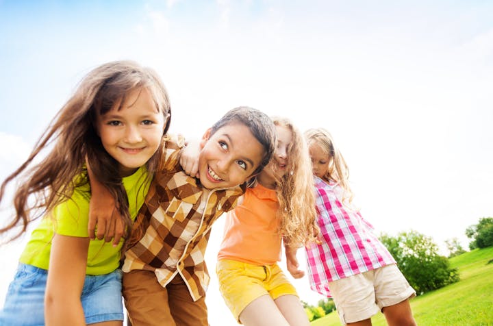 Four children smiling and hugging outdoors on a sunny day with a grassy background.