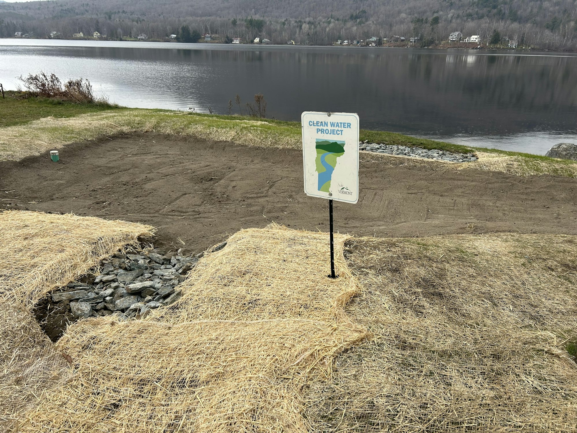 A sign for a "Clean Water Project" near a body of water, with straw-covered ground and rocks.