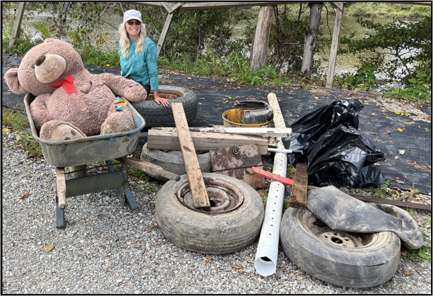 A person with a wheelbarrow holding a large teddy bear, old tires, wood, a black bag, and assorted junk outside.