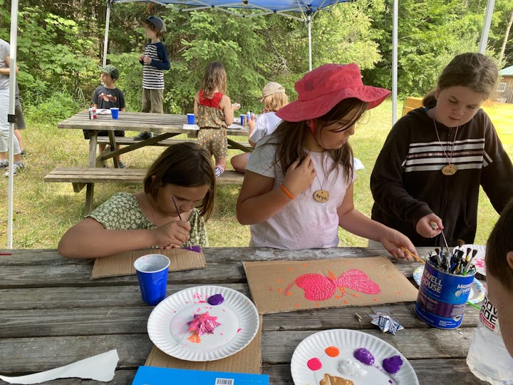 Kids are painting on cardboard in an outdoor setting, with art supplies and drinks on tables, enjoying a creative activity.