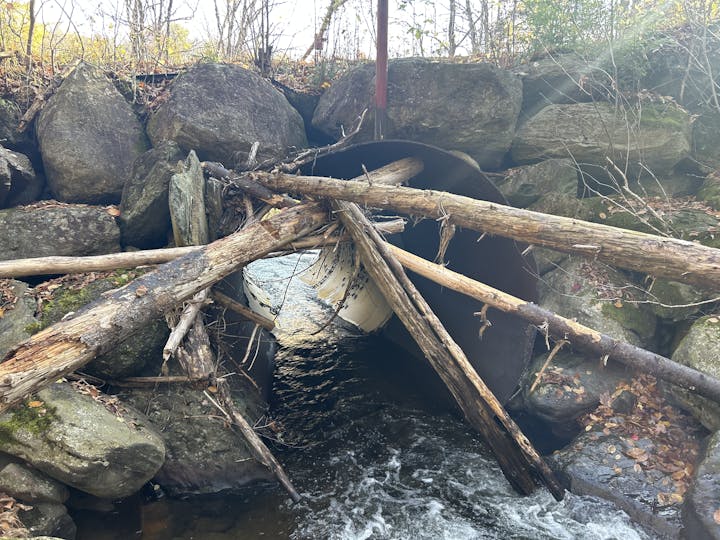 A culvert blocked by fallen logs and branches, surrounded by large rocks and flowing water beneath.