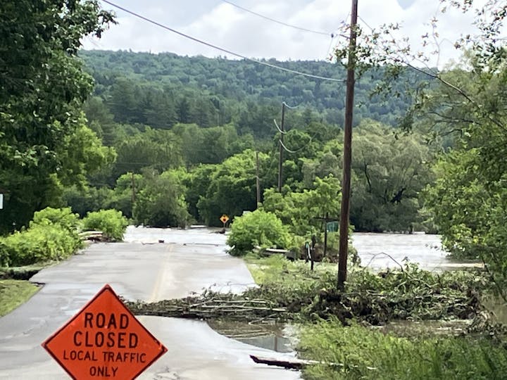 Flooded road with "ROAD CLOSED" sign, debris, trees, and hills in the background.