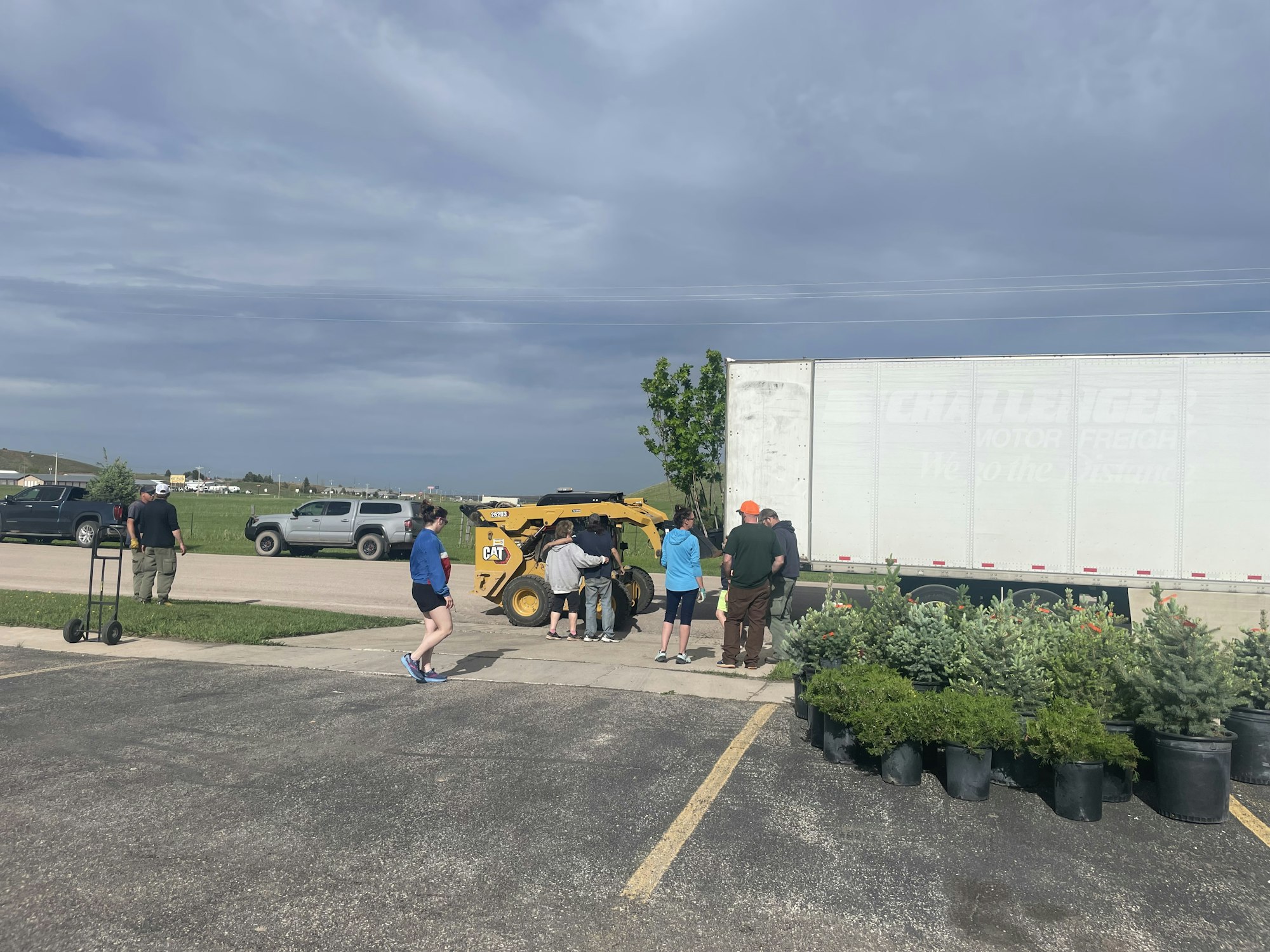 People are gathered around a small yellow construction vehicle near a truck; plants in pots are on the pavement.