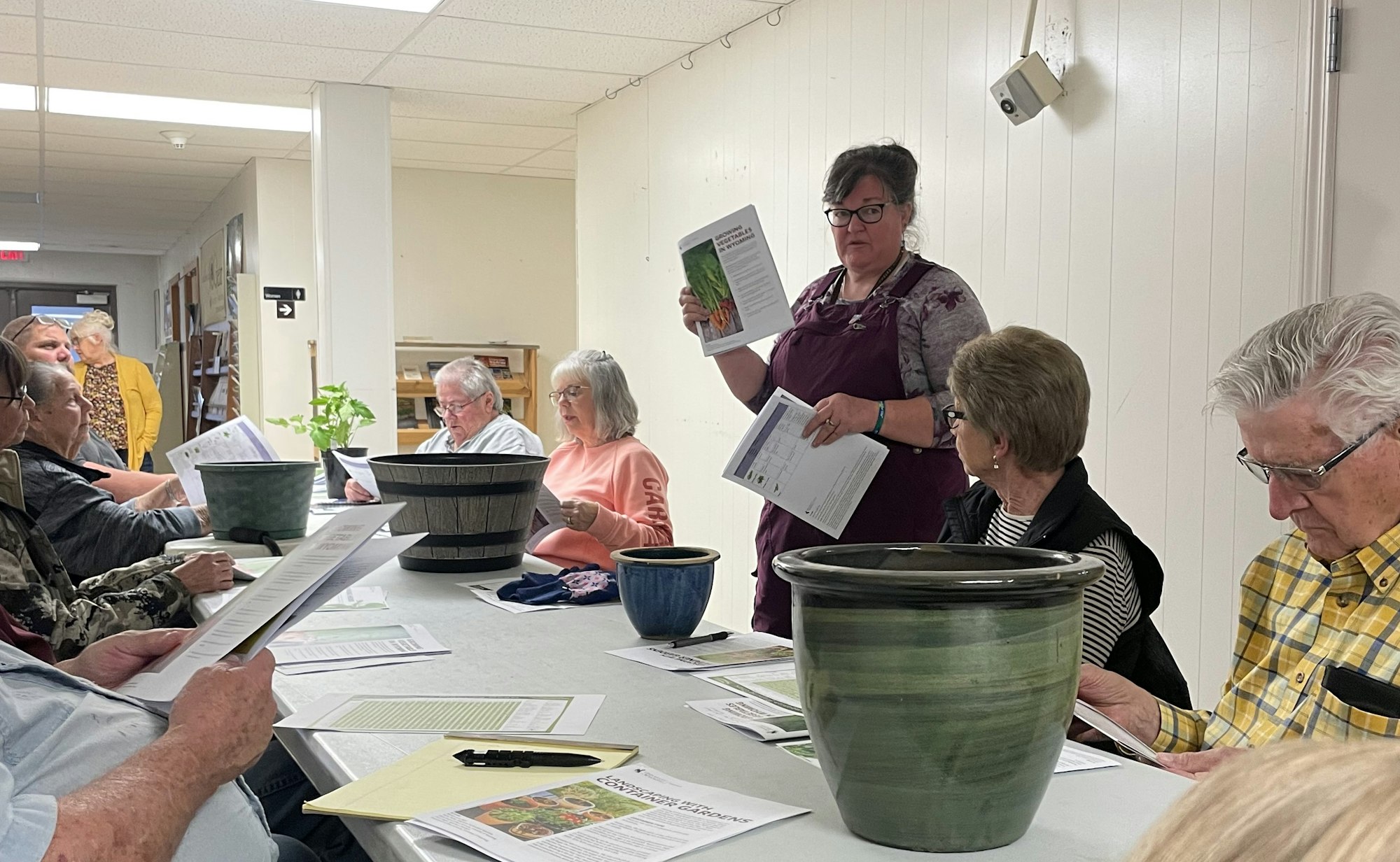 Group of adults at a meeting with papers and flower pots, one person presenting.