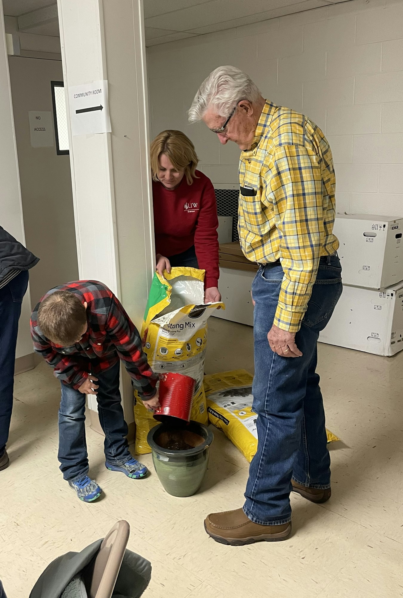 Three people, possibly volunteers, are pouring and measuring contents from bags into a bucket inside a room.
