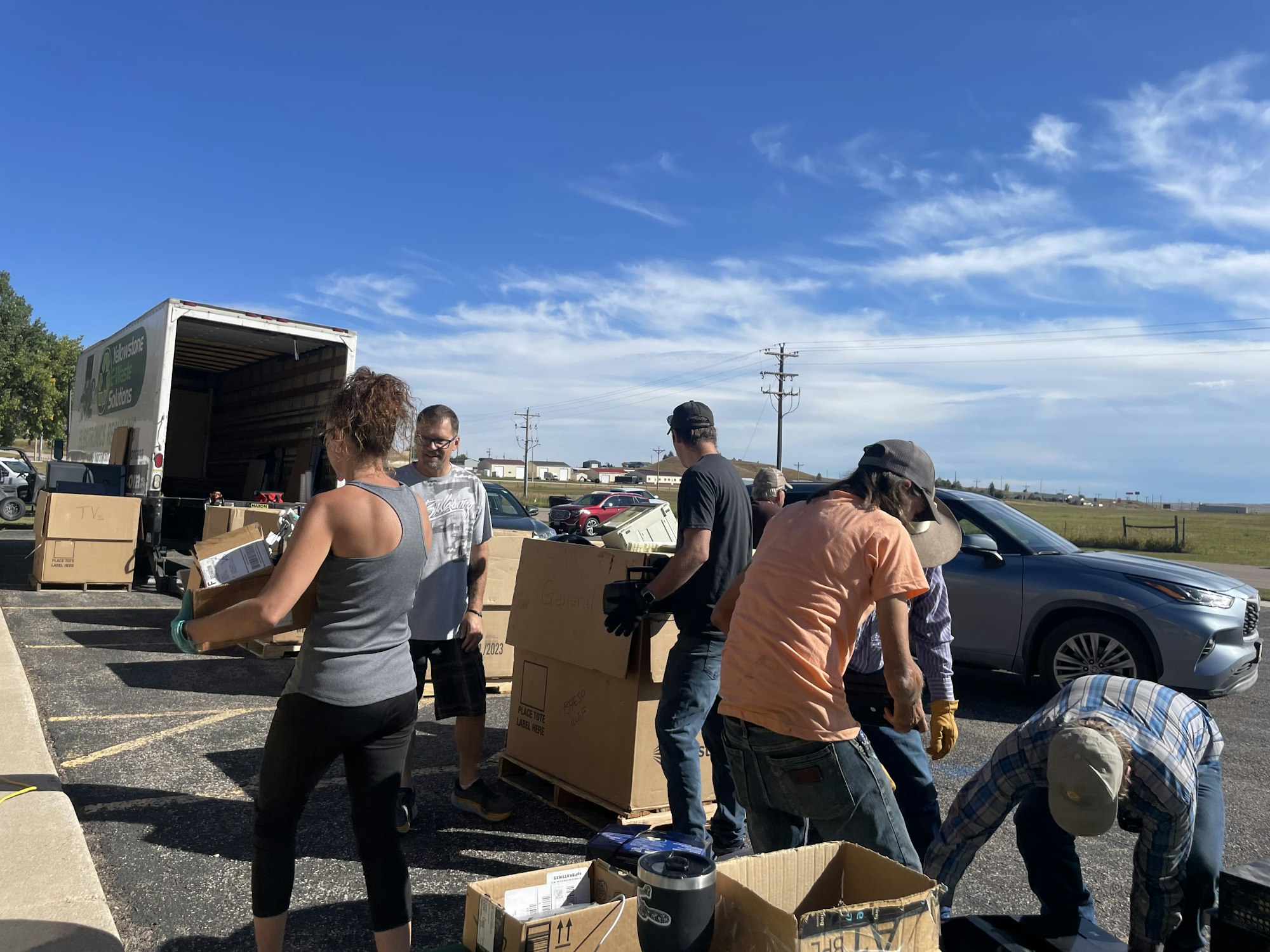 People unloading electronics from vehicles in a parking lot under a blue sky.