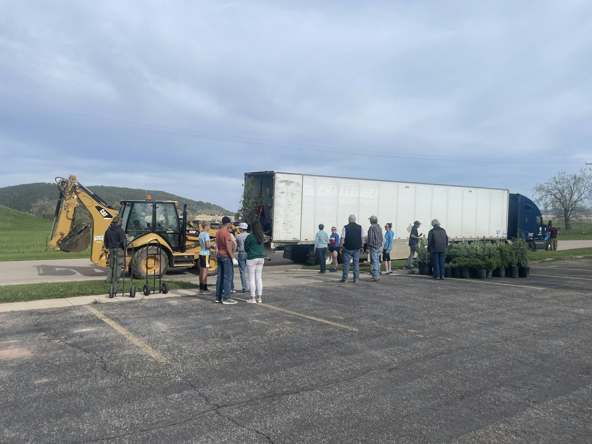 A group of people stand near a truck, next to potted plants and a backhoe, in an outdoor setting.