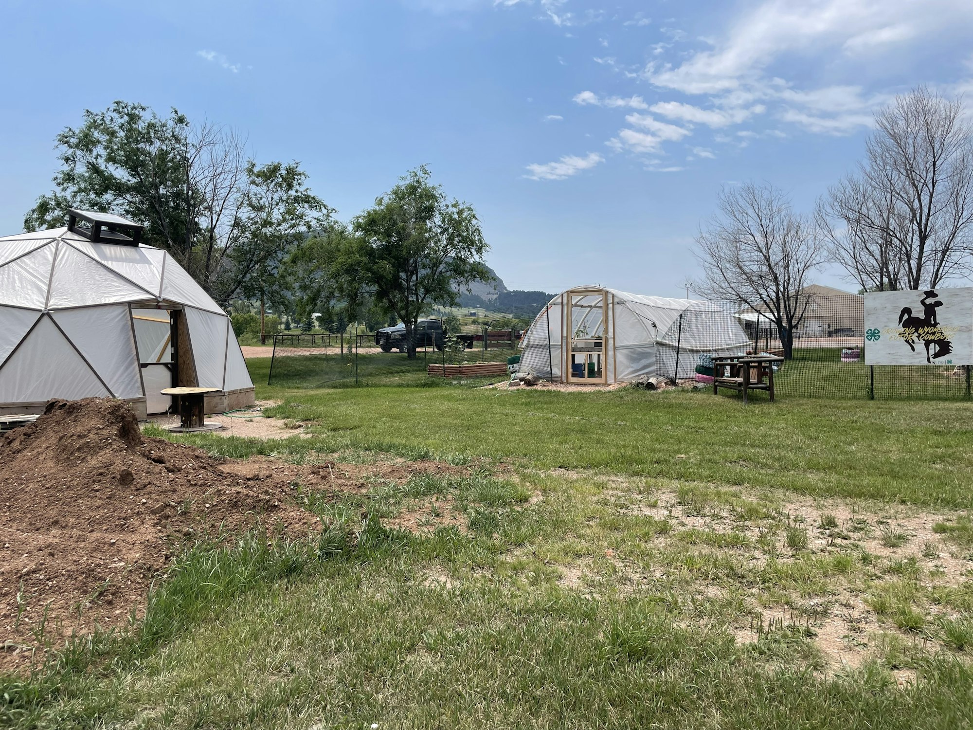 Geodesic dome and greenhouse with sign, bench, and trees against a clear sky.