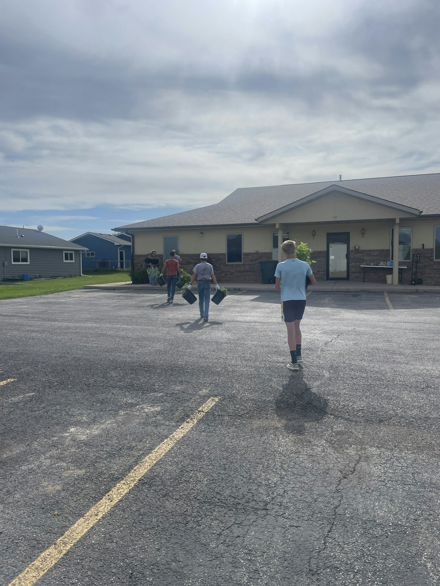 People walking and carrying objects near a building under a cloudy sky.