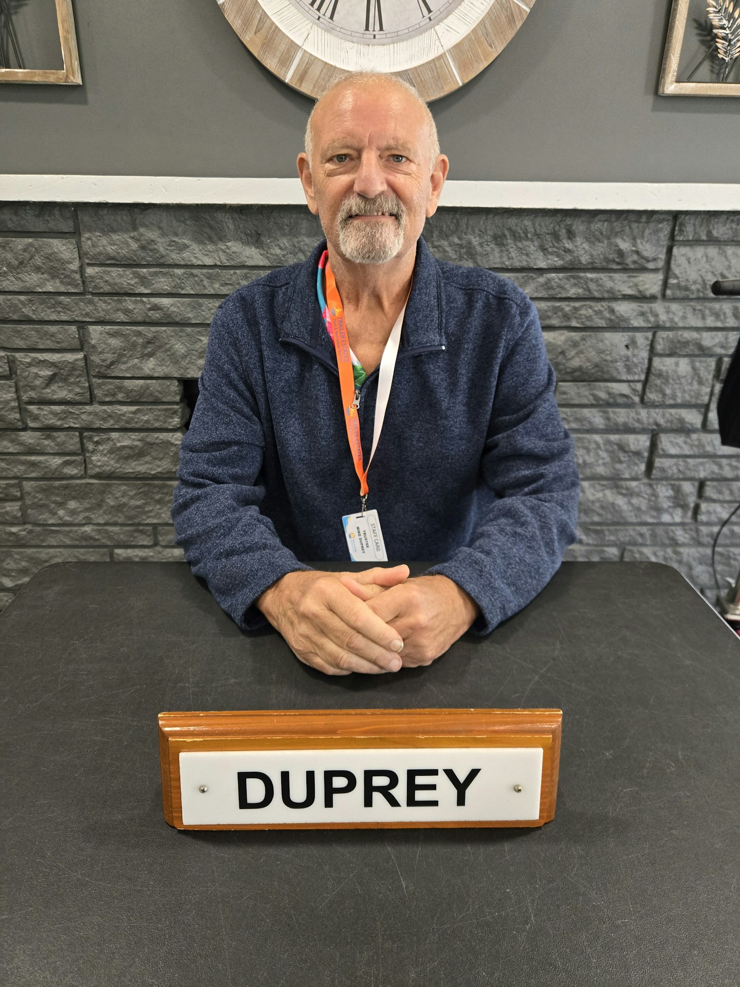 An older man in a blue sweater sits at a desk with a nameplate reading "DUPREY" and a staff ID badge around his neck.