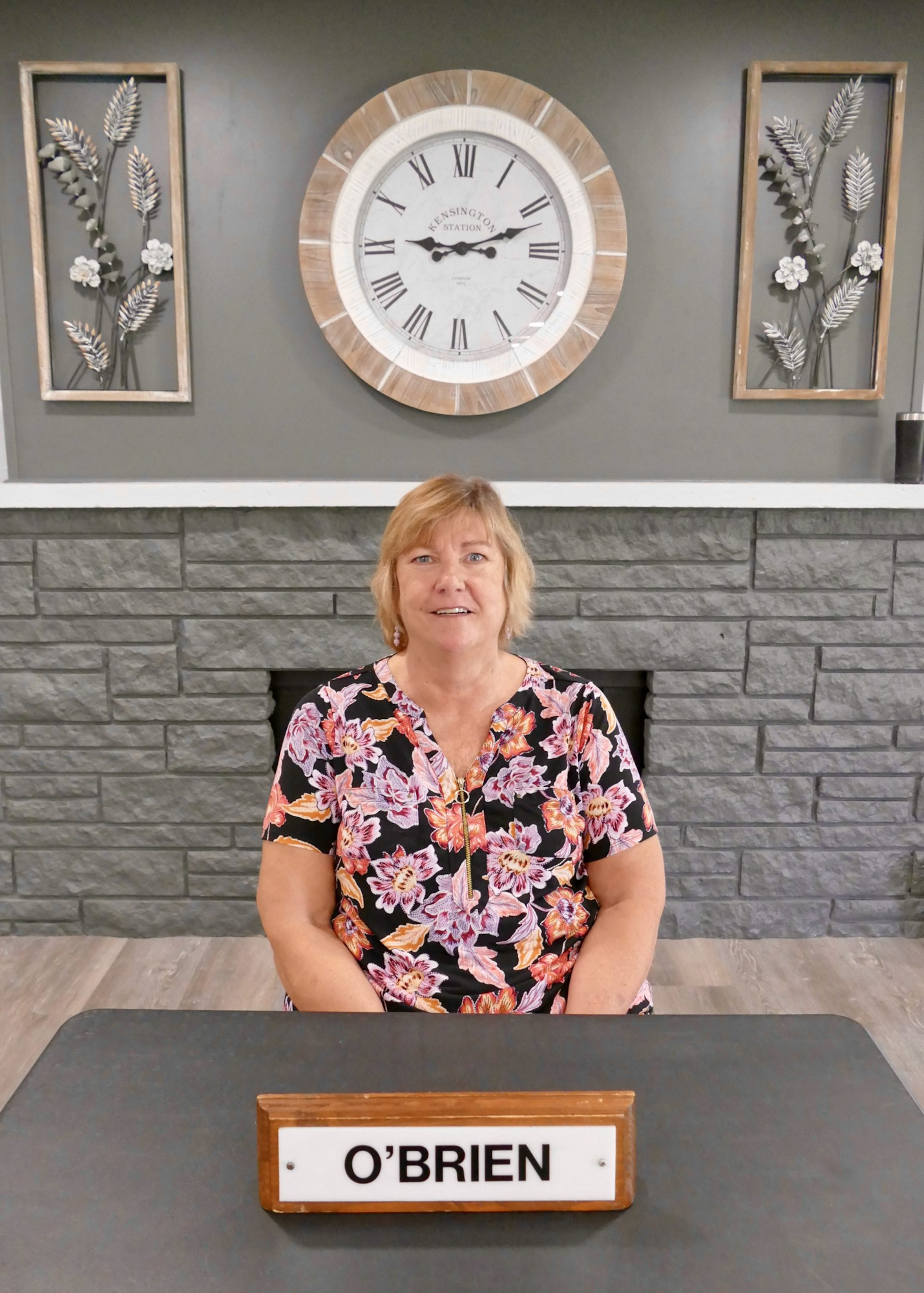 A woman with blonde hair is sitting at a table with an "O'BRIEN" nameplate, in front of a decorative wall with a clock.