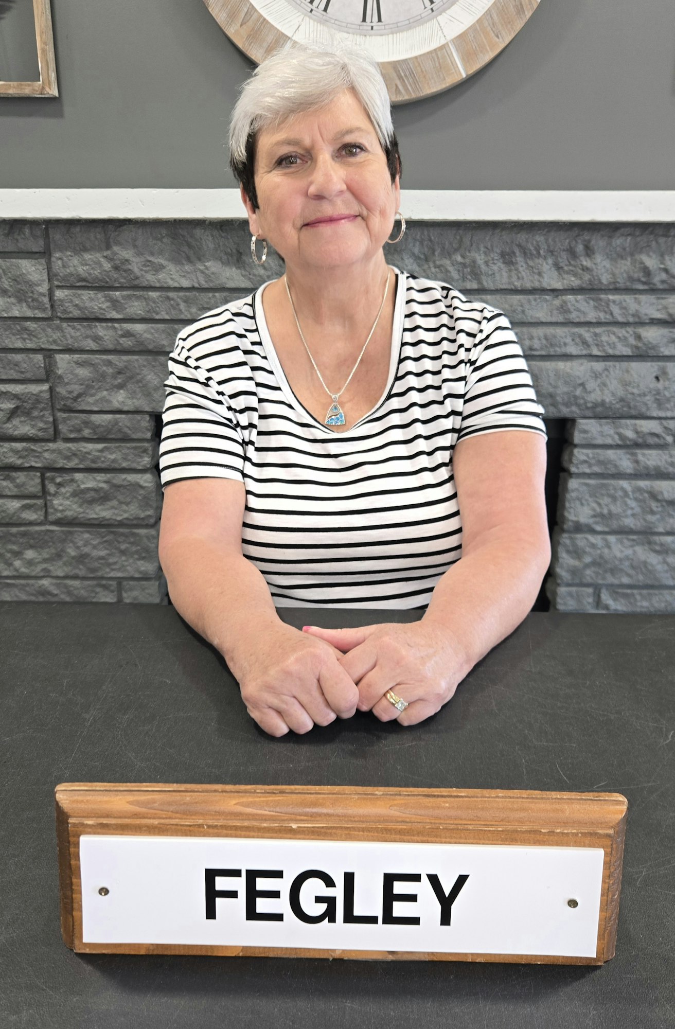 A person seated at a table with a nameplate reading "FEGLEY"; wearing a striped shirt and necklace.