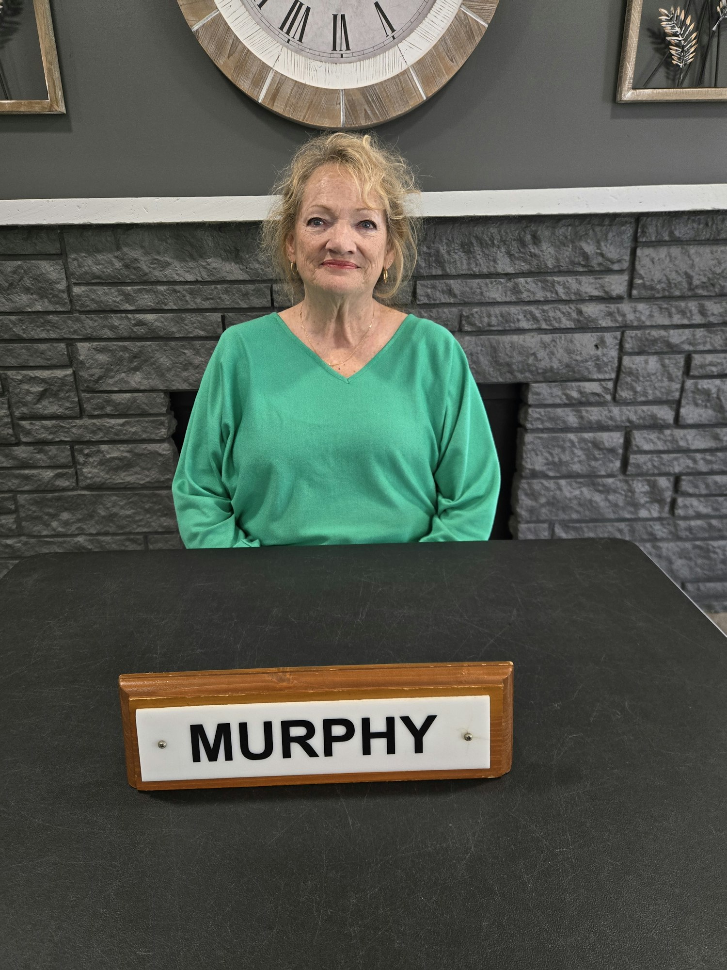 A woman in a green shirt smiles beside a nameplate that reads "MURPHY" against a textured wall with décor.