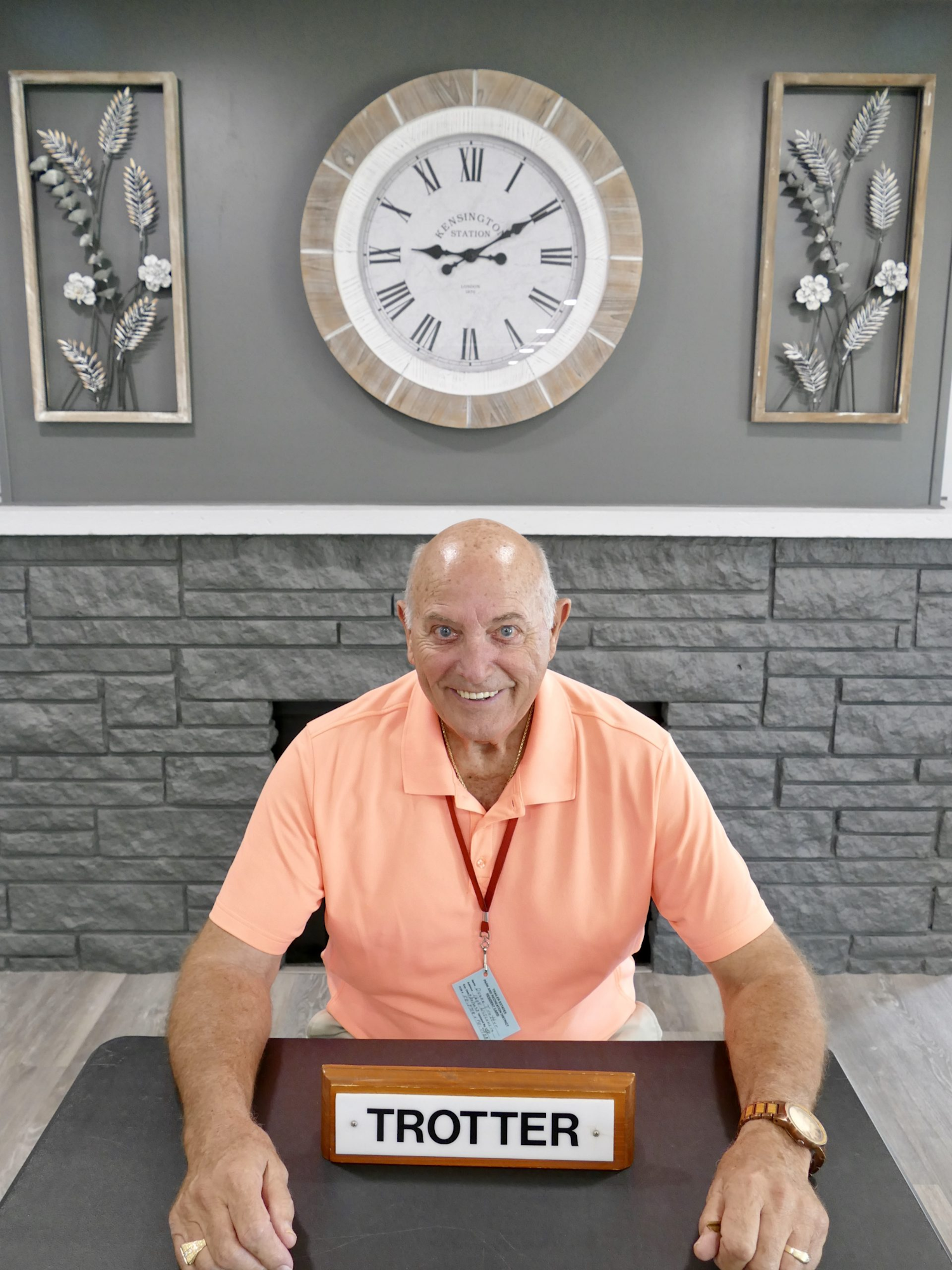 A smiling man in a pink shirt sits at a table with a nameplate "TROTTER." Behind him is a decorative clock and wall art.