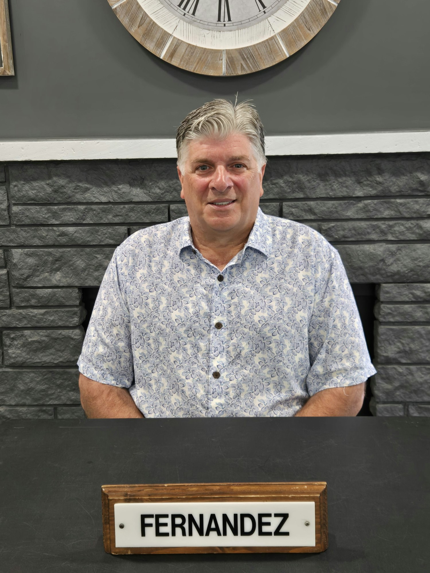 A person in a patterned shirt stands next to a sign with "FERNANDEZ" on it; a clock is on the wall.