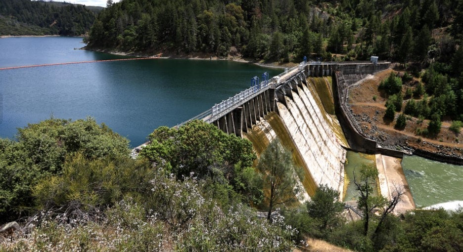 A dam with water flowing over it, surrounded by a forested landscape and a body of water on one side.