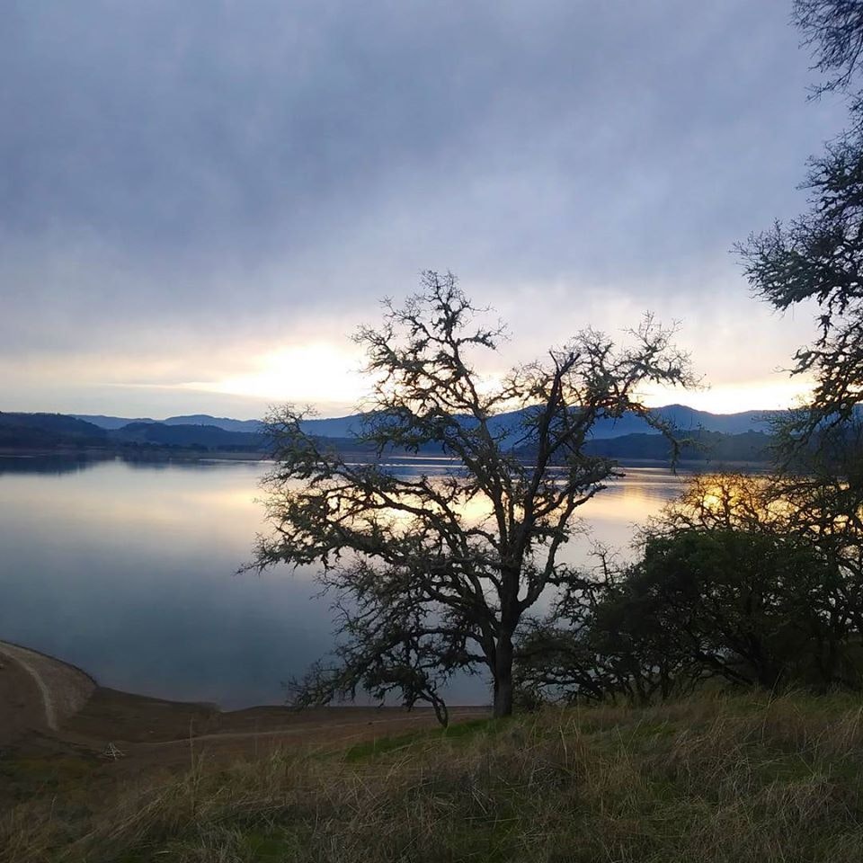 A serene landscape featuring a lake, trees, mountains, and a cloudy sky at sunset, reflecting calm waters.