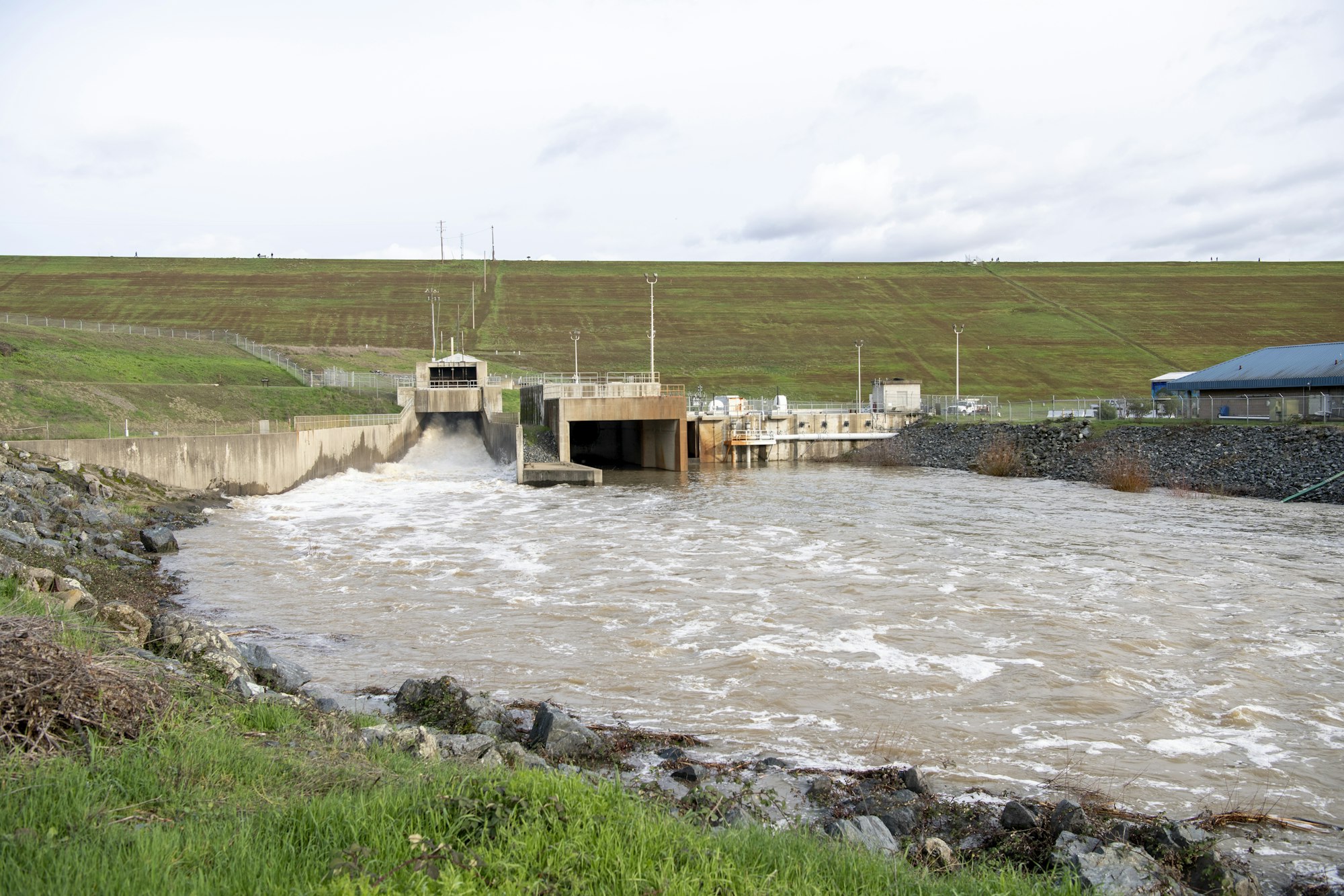 The image shows a water control infrastructure, with a flow of water, a dam structure, and green hills in the background.