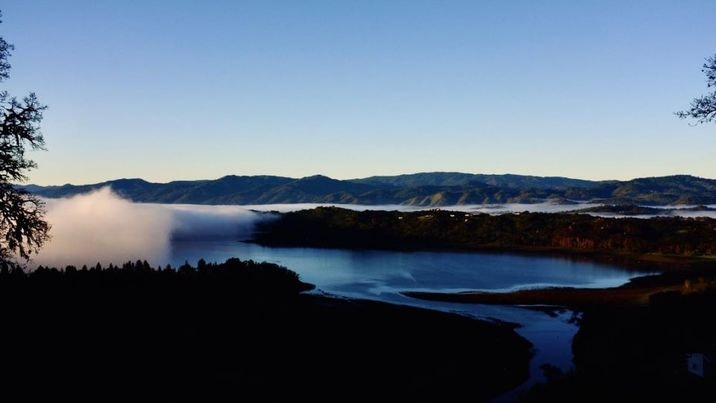 A serene landscape featuring a lake surrounded by mountains and fog, under a clear blue sky.