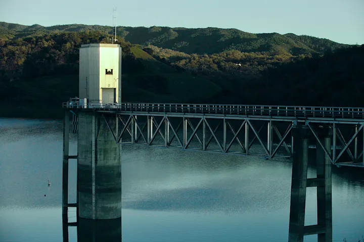 The image shows a water reservoir tower with a bridge extending over a calm body of water surrounded by hills.