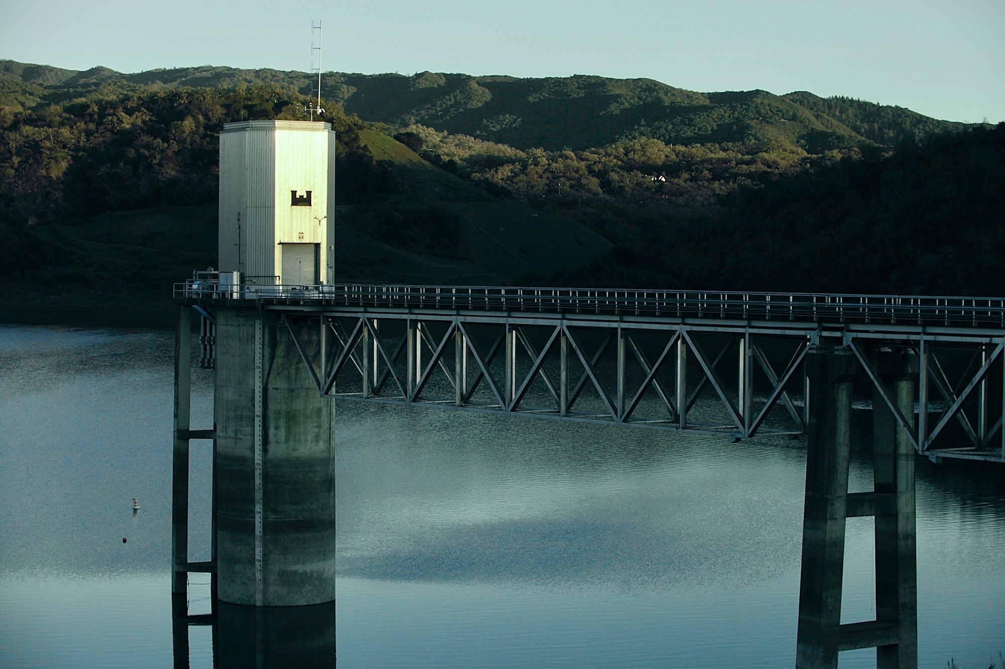 The image shows a water reservoir tower with a bridge extending over a calm body of water surrounded by hills.